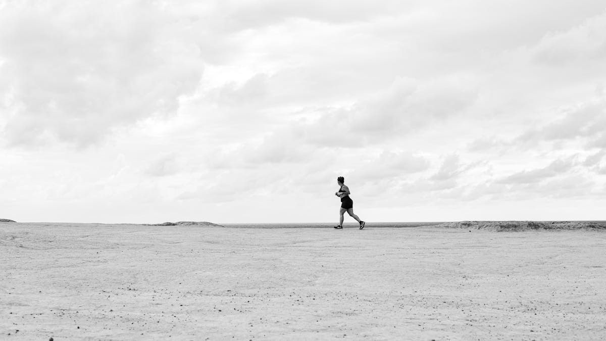 A lone runner traverses a vast, empty Brazilian beach under dramatic cloudy skies in black and white.