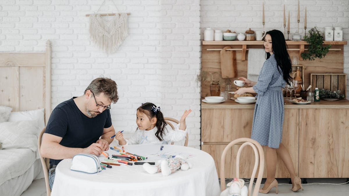 Father and daughter bond over crafts while mother prepares food in a cozy home setting.